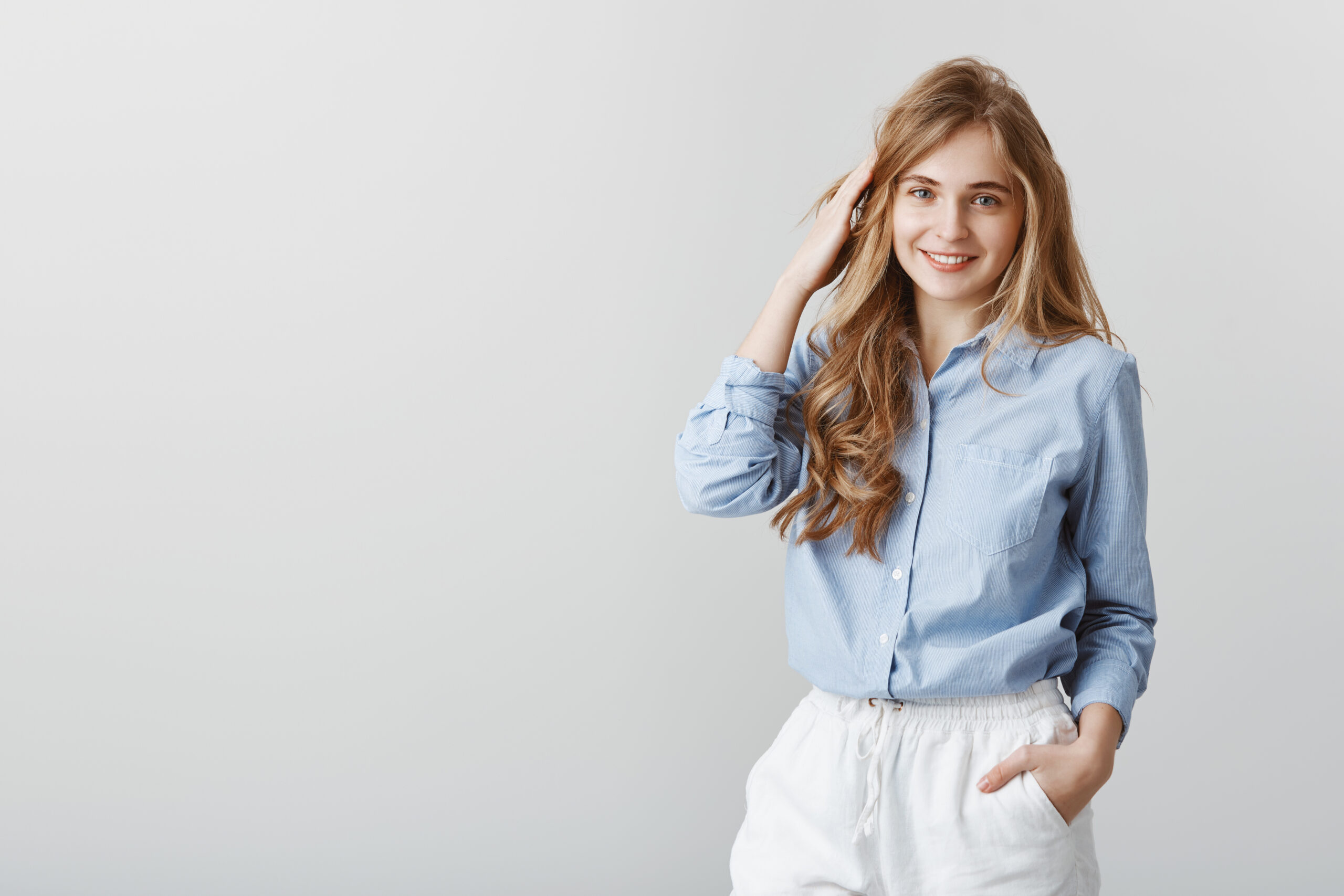 Girl having ordinary chit-chat with friends. Studio shot of charming young caucasian female blonde in blue blouse, touching hair and smiling cheerfully, being polite with client during work in office з днем народження Марина