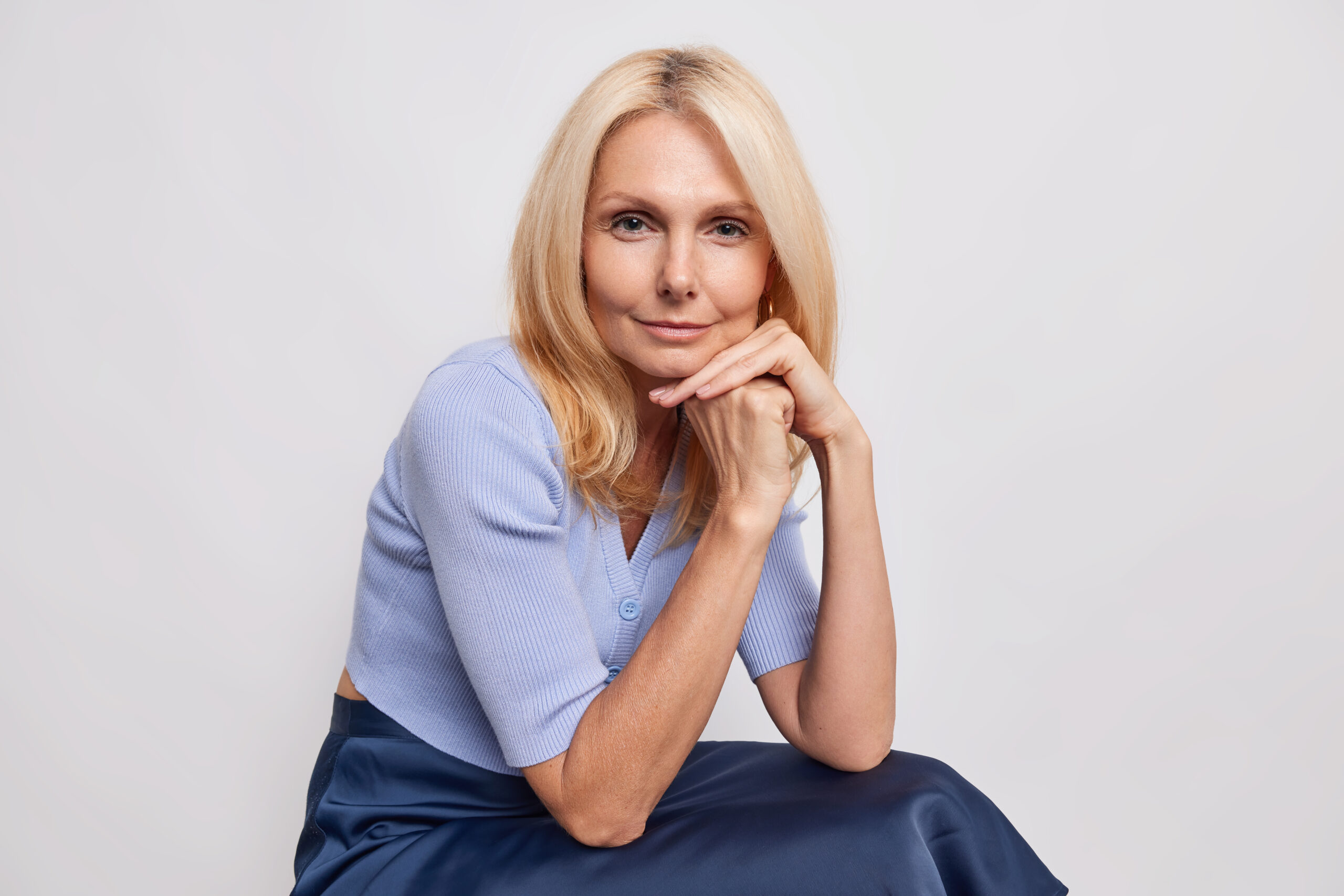 Portrait of self confident forty years old woman with blonde hair keeps hands near face looks directly at camera wears blue jumper and skirt sits against white background thinks about something з днем народження Віка своїми словами